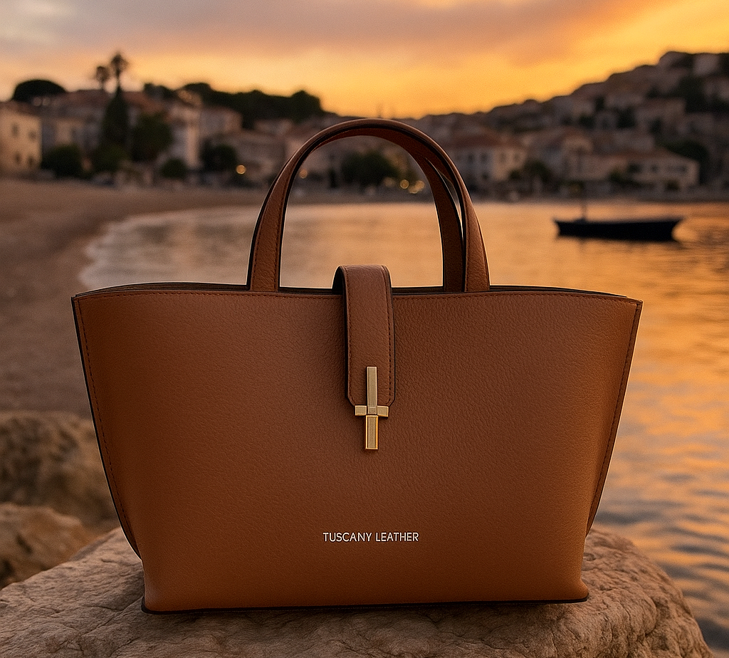 Caramel Tuscany Leather handbag resting on a seaside rock at sunset, with a soft golden sky and coastal village in the background