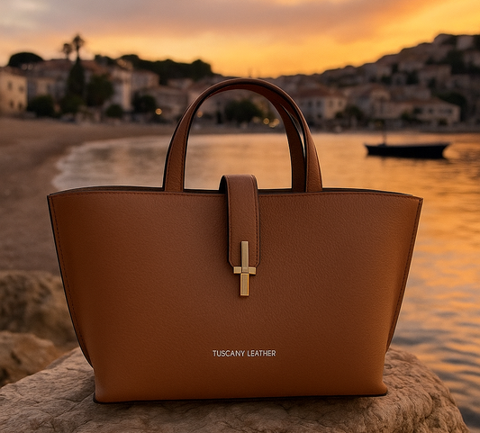 Caramel Tuscany Leather handbag resting on a seaside rock at sunset, with a soft golden sky and coastal village in the background