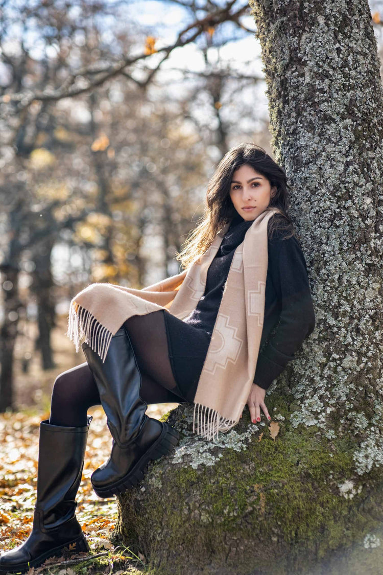 Woman wearing a beige alpaca wool scarf, sitting against a tree in a peaceful autumn forest, styled with black boots and knitwear.