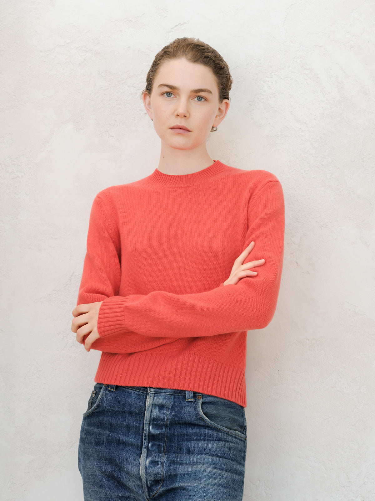 A woman wearing a coral-red knit crewneck sweater and blue jeans, standing with arms crossed against a textured off-white wall.