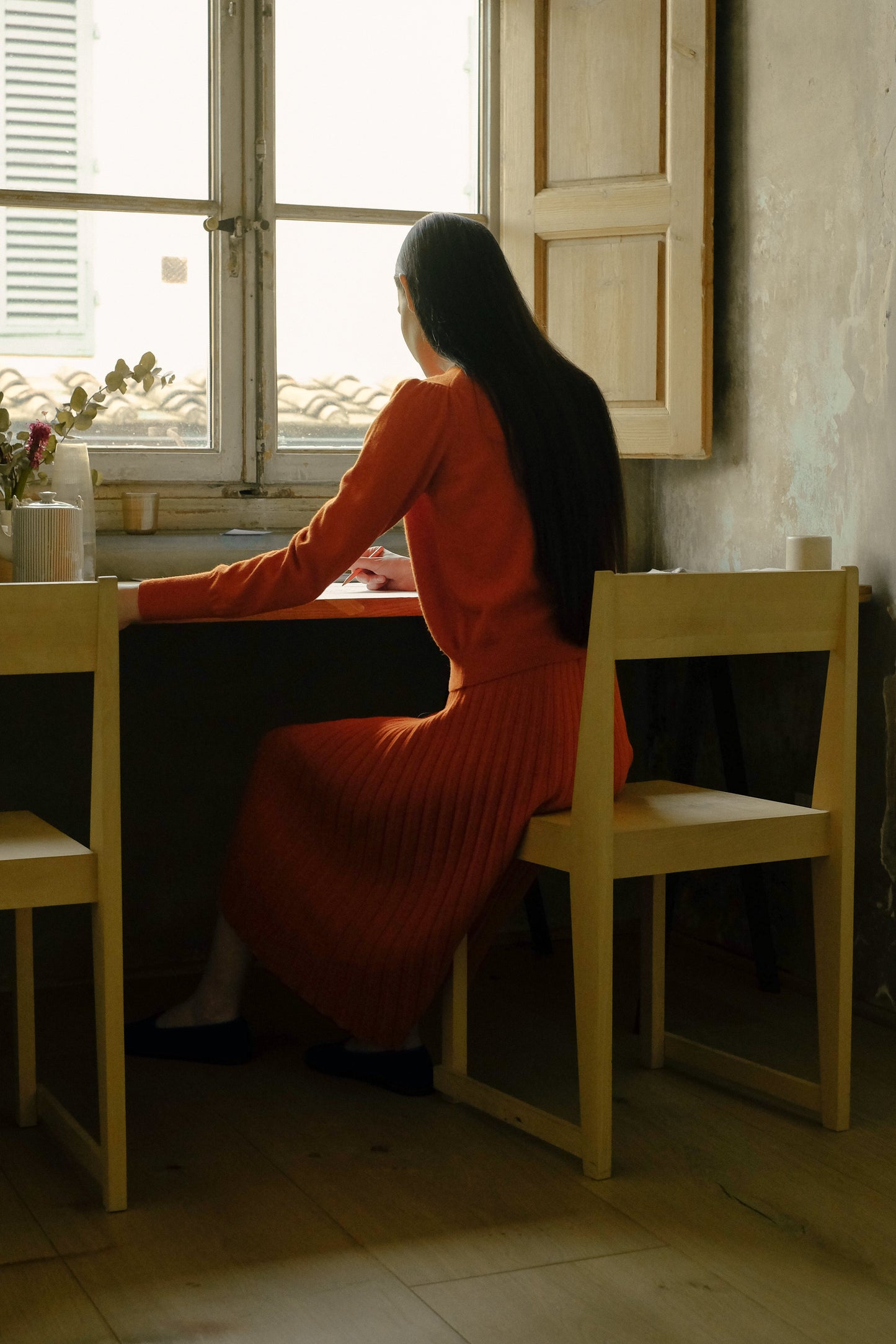 Woman in an orange pleated knit skirt and orange sweater sitting at a table by a window in a dimly lit room.