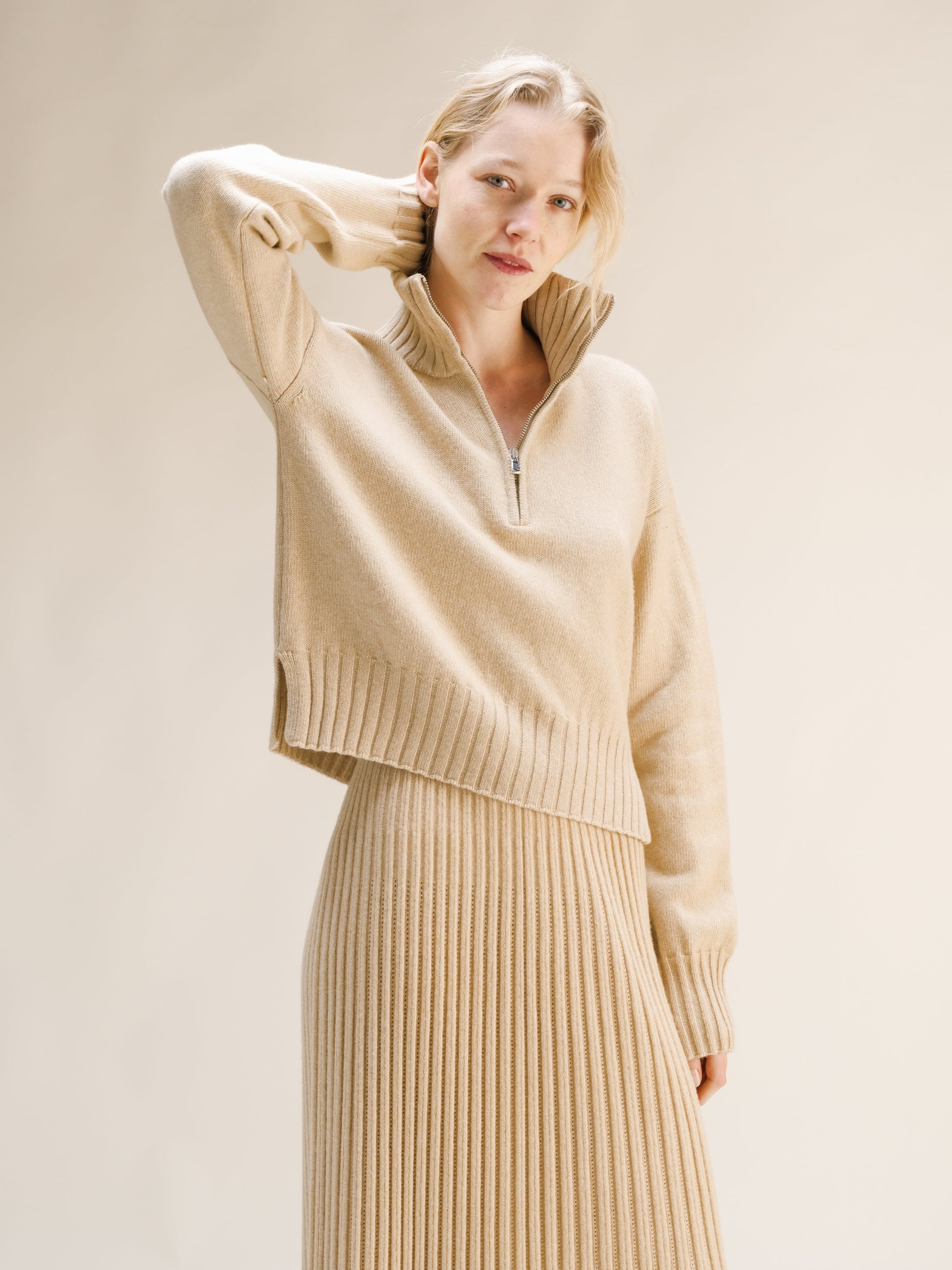 Woman wearing a beige sweater and pleated skirt against a plain background