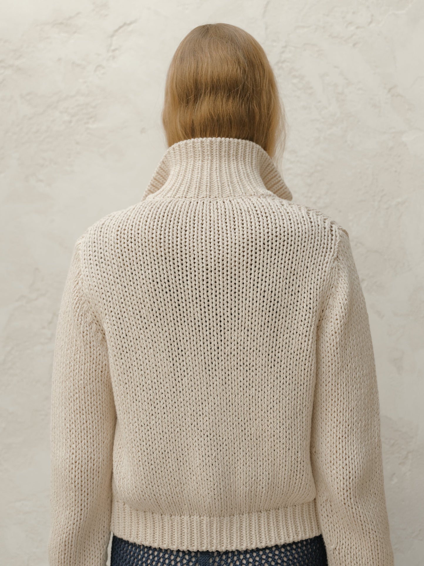 Back view of a model wearing a cream chunky knit bomber jacket with a ribbed hem against a textured light wall.