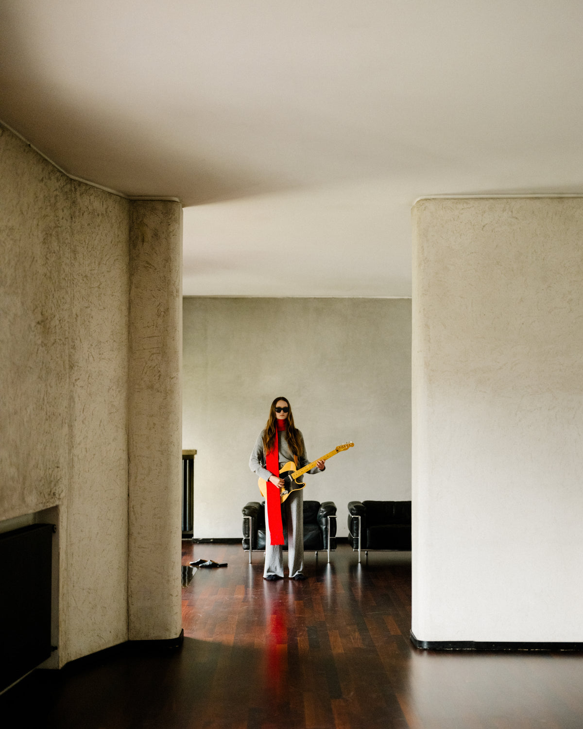 Person wearing grey cashmere mix pull col rond with a red scarf and sunglasses holding an electric guitar in a minimalist living room.