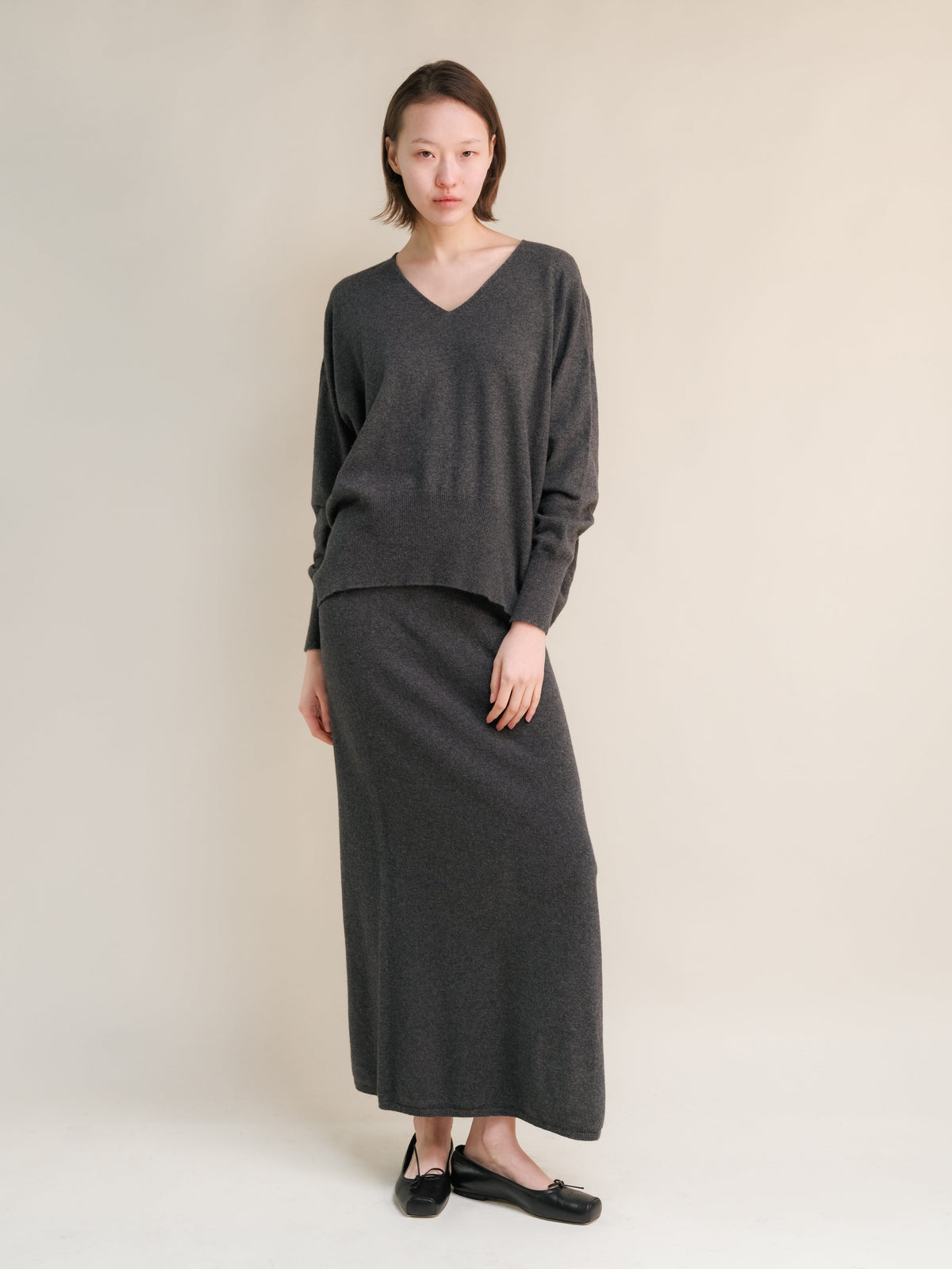 Model trägt Anthrazitgrau V-Ausschnitt-Strickpullover aus Kaschmirmischung with a matching high waist langer Rock in a minimalist studio.