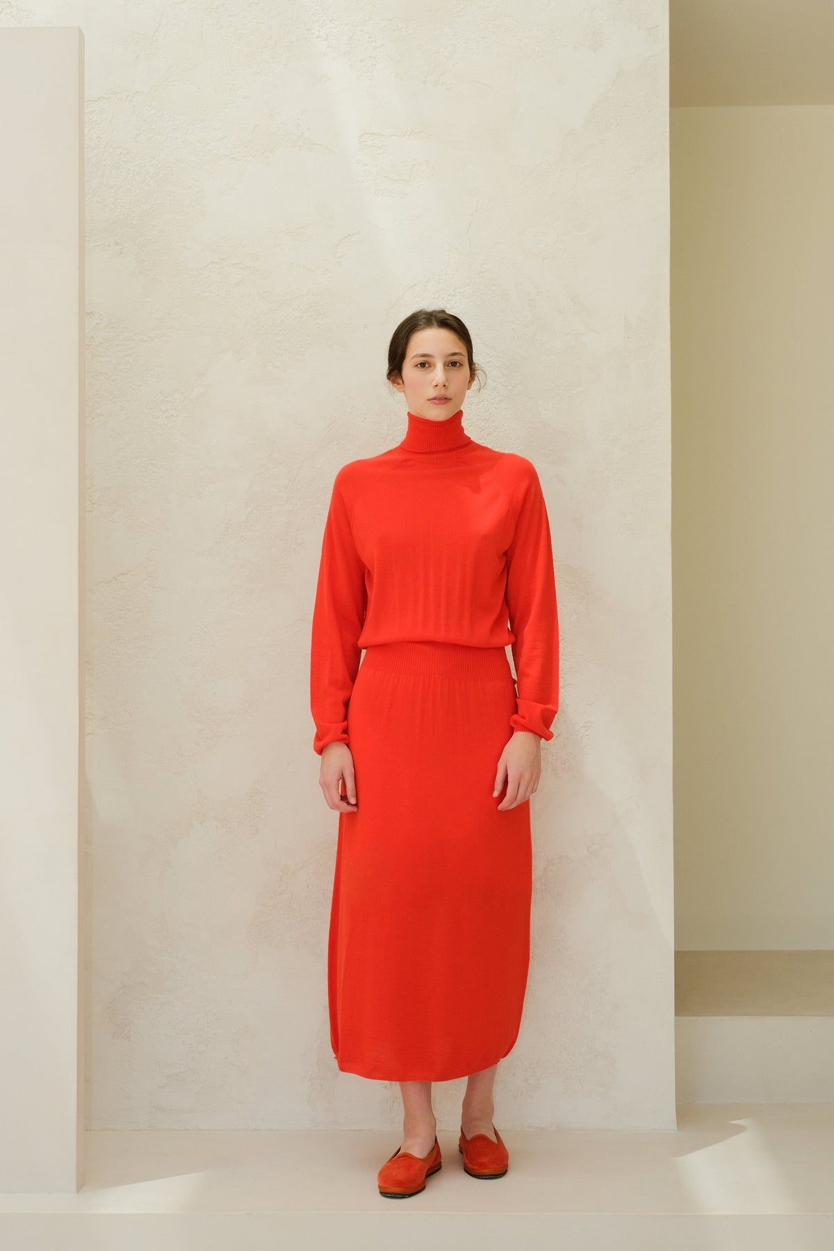 Model facing forward in a felrood 3D knit merino wool dress with a hoge kraag and geribde taille, midi-length.