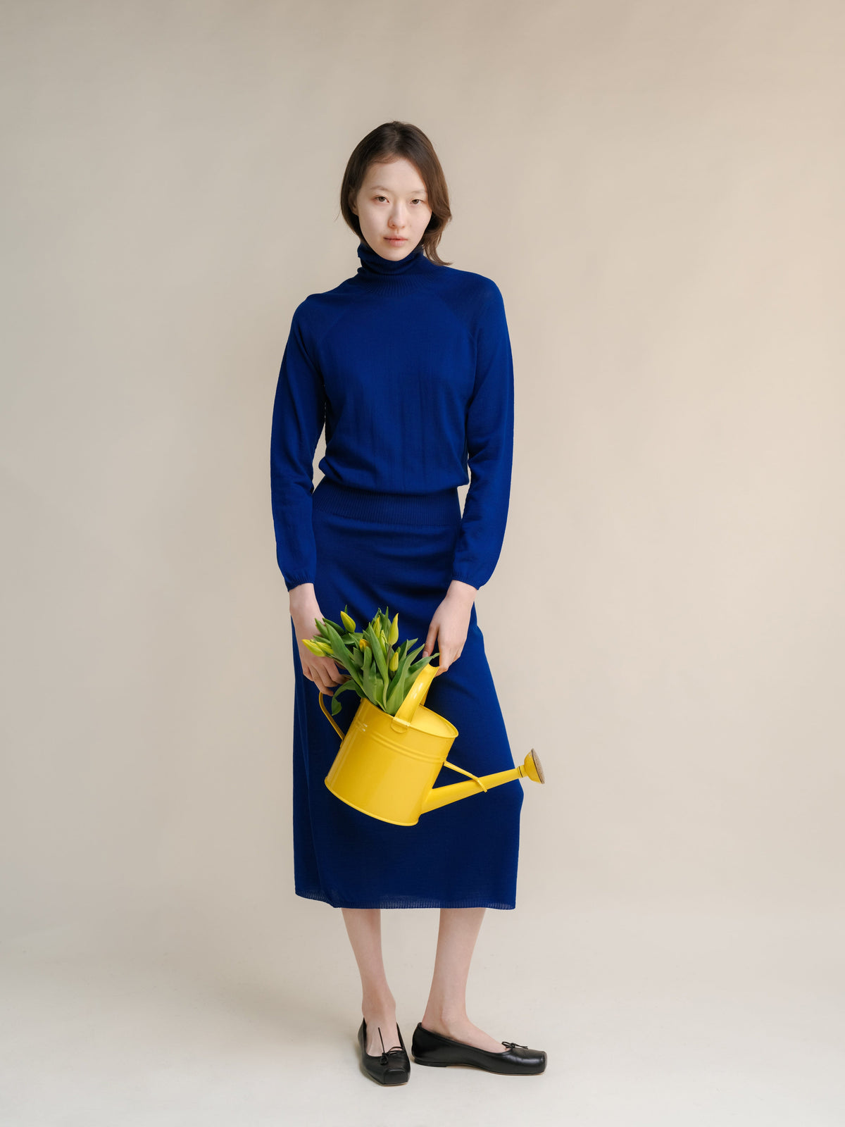 Model in a royal blue coltrui dress met een gele gieter with green plants against a neutral backdrop