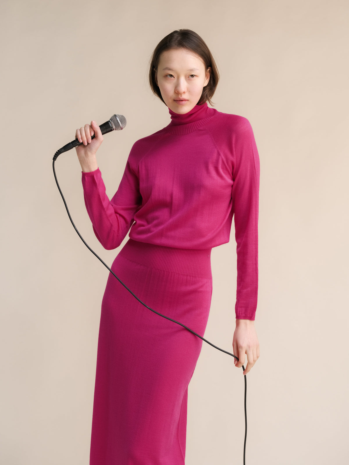 Model draagt magenta 3D knit merino wool dress posing with a microphone in a studio