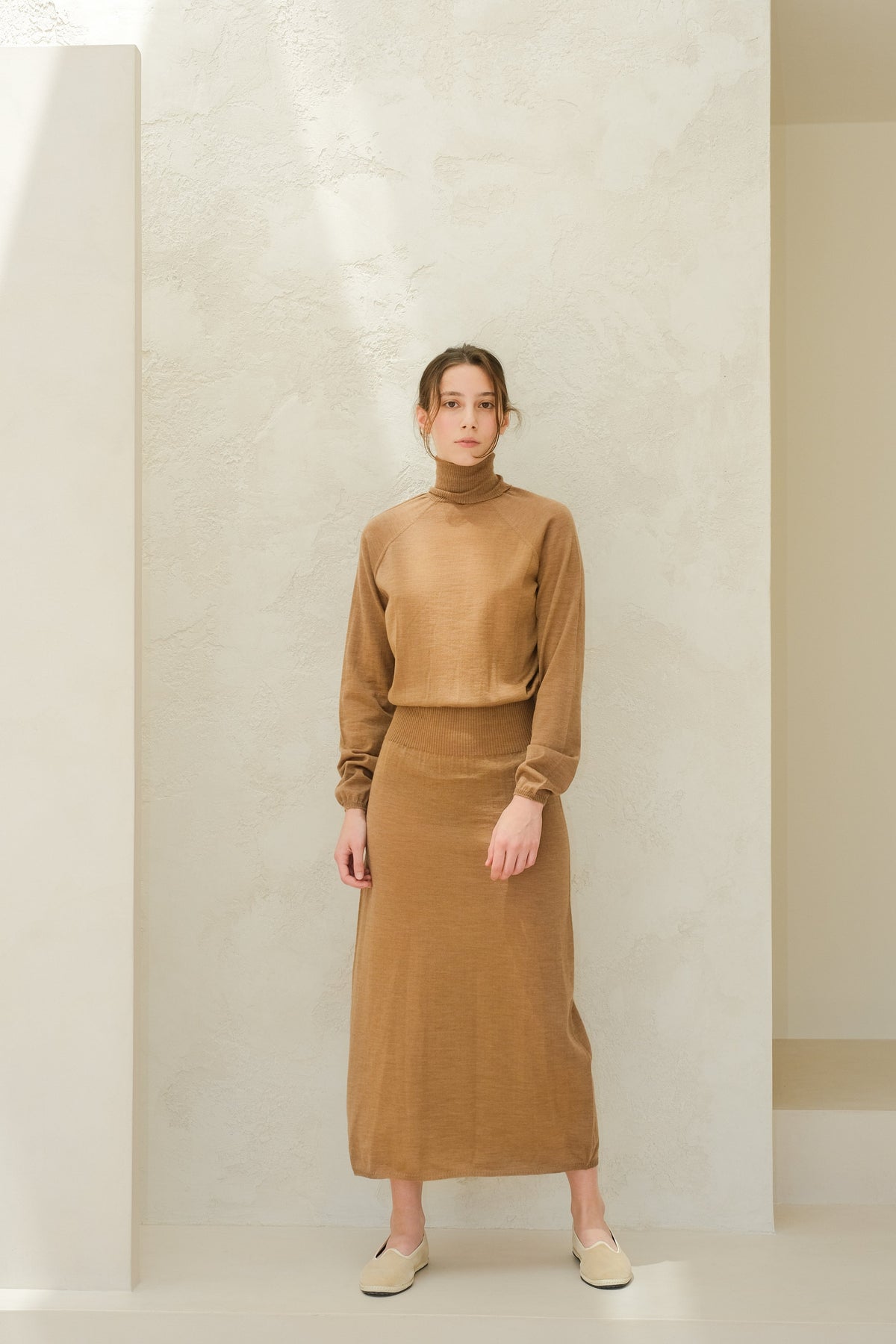 Full-body view of a model in camel brown 3D knit merino dress set with a coltrui top and ribbed-waist skirt, beige slip-on shoes