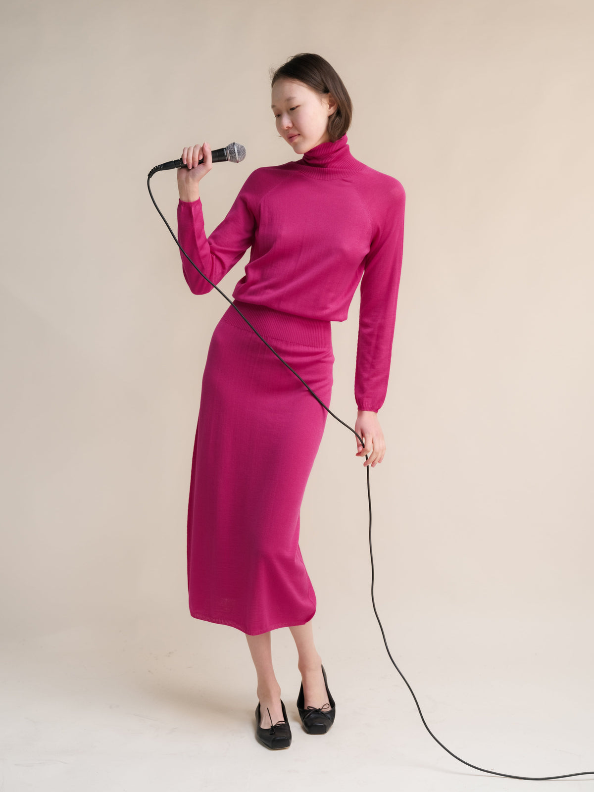 Model draagt magenta 3D-Knit jurk van extra fijne merinowol with a coltrui, met een microfoon.