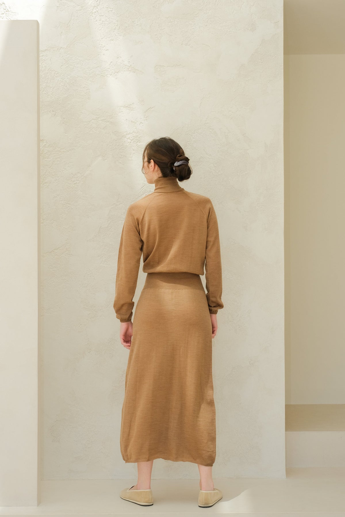 Back view of a model in camel brown knit dress with bijpassende lange rok, hair in a bun.