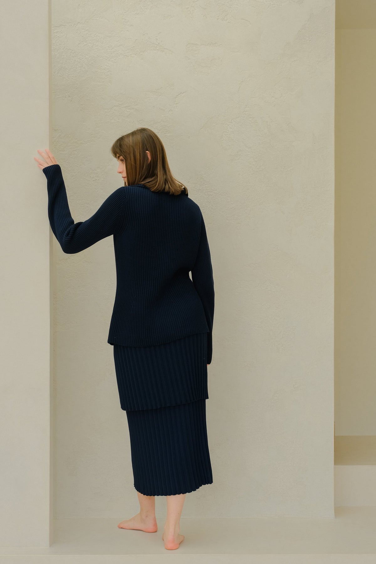 Back view of a mannequin porcognact a bleu marine knit plissé skirt set against a beige wall.