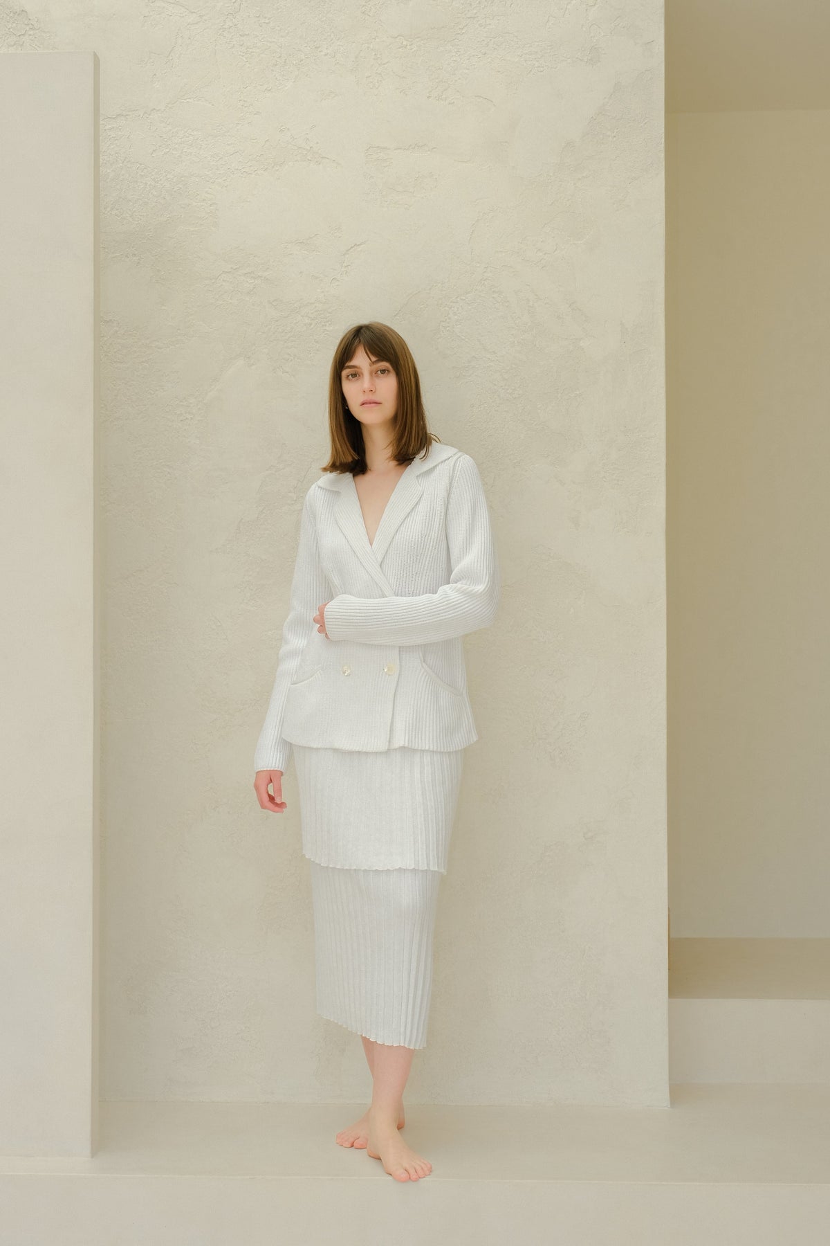 Vue latérale of the model in a pale gray blazer en maille and jupe longue plissée assortie against a textured light wall.
