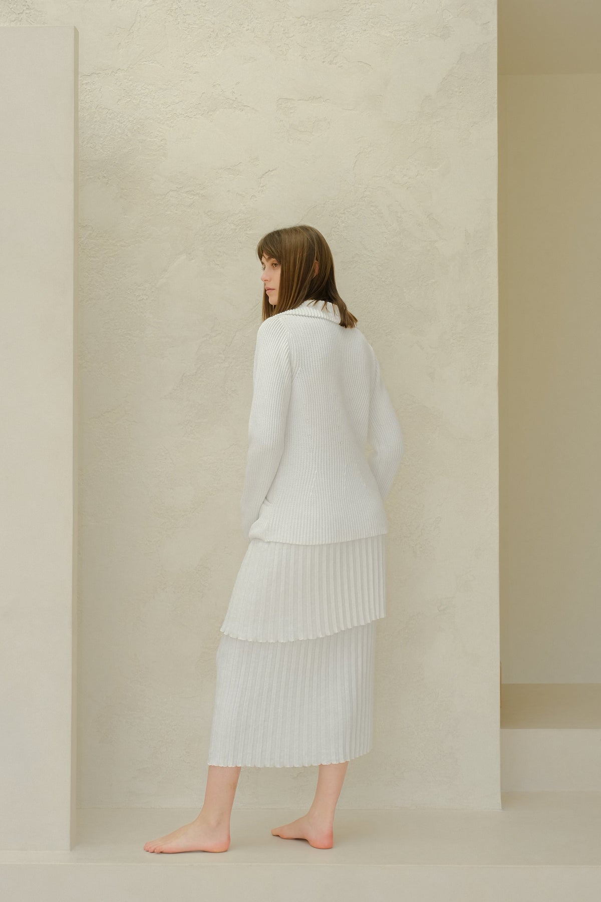 Vue latérale of a mannequin porcognact a white fine cotton plissé knit skirt with a pull assorti against a textured beige wall.