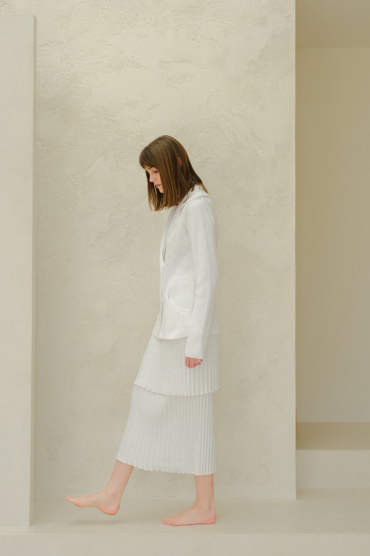 Side profile of a model in a white blazer en maille with a layered jupe longue plissée from Fine Cotton Plissé