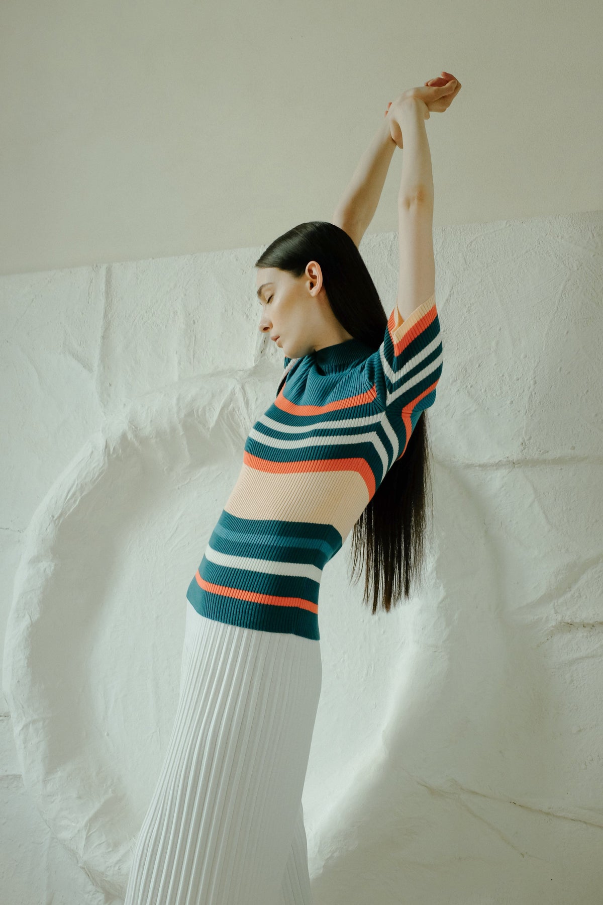 Profile of a model in a multicolor striped knit top and a white jupe longue plissée, arms raised overhead.