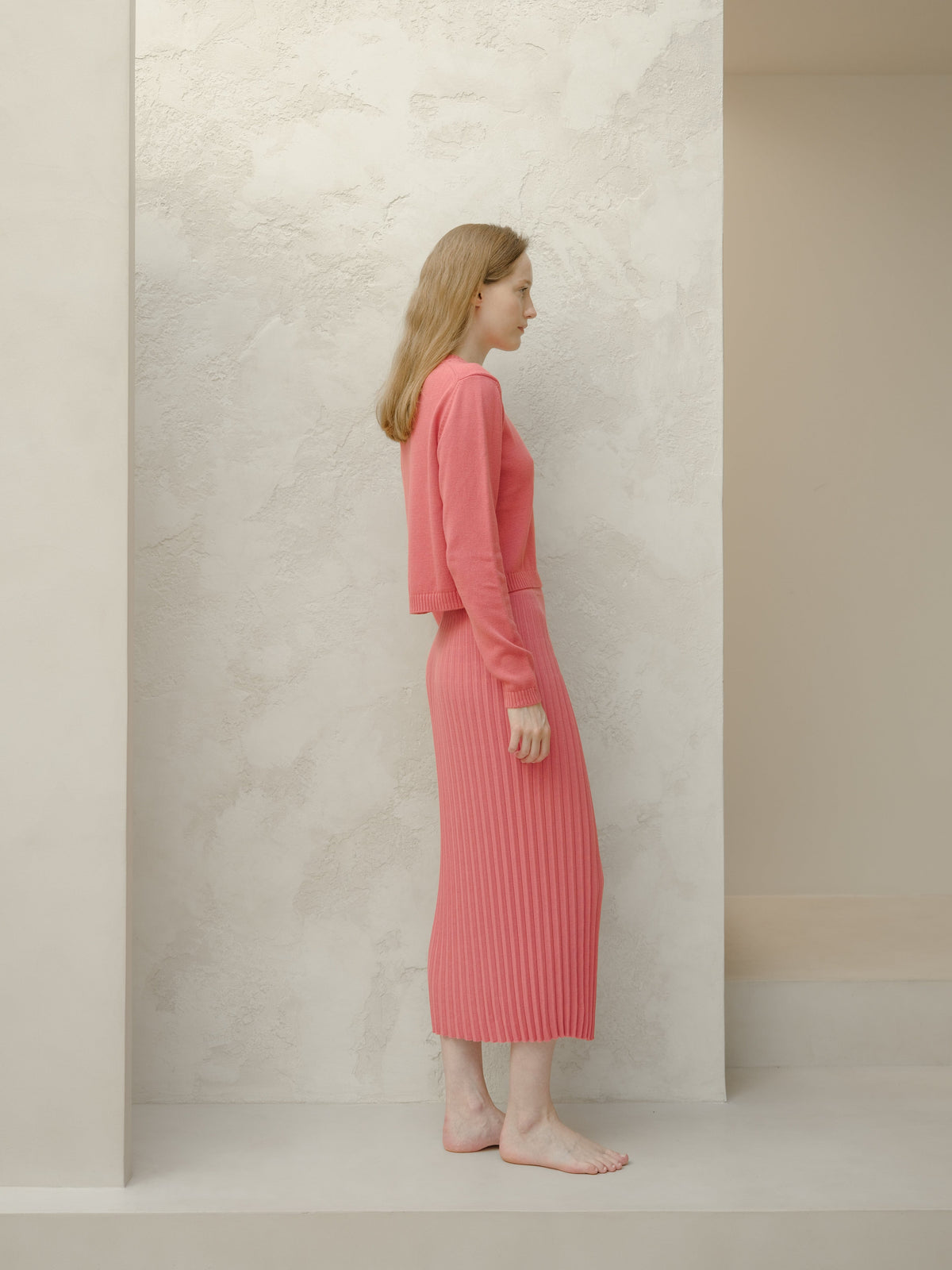 Vue latérale of a model in a rose poudré 3D Knit jupe longue plissée en coton fin with a cropped pull assorti.