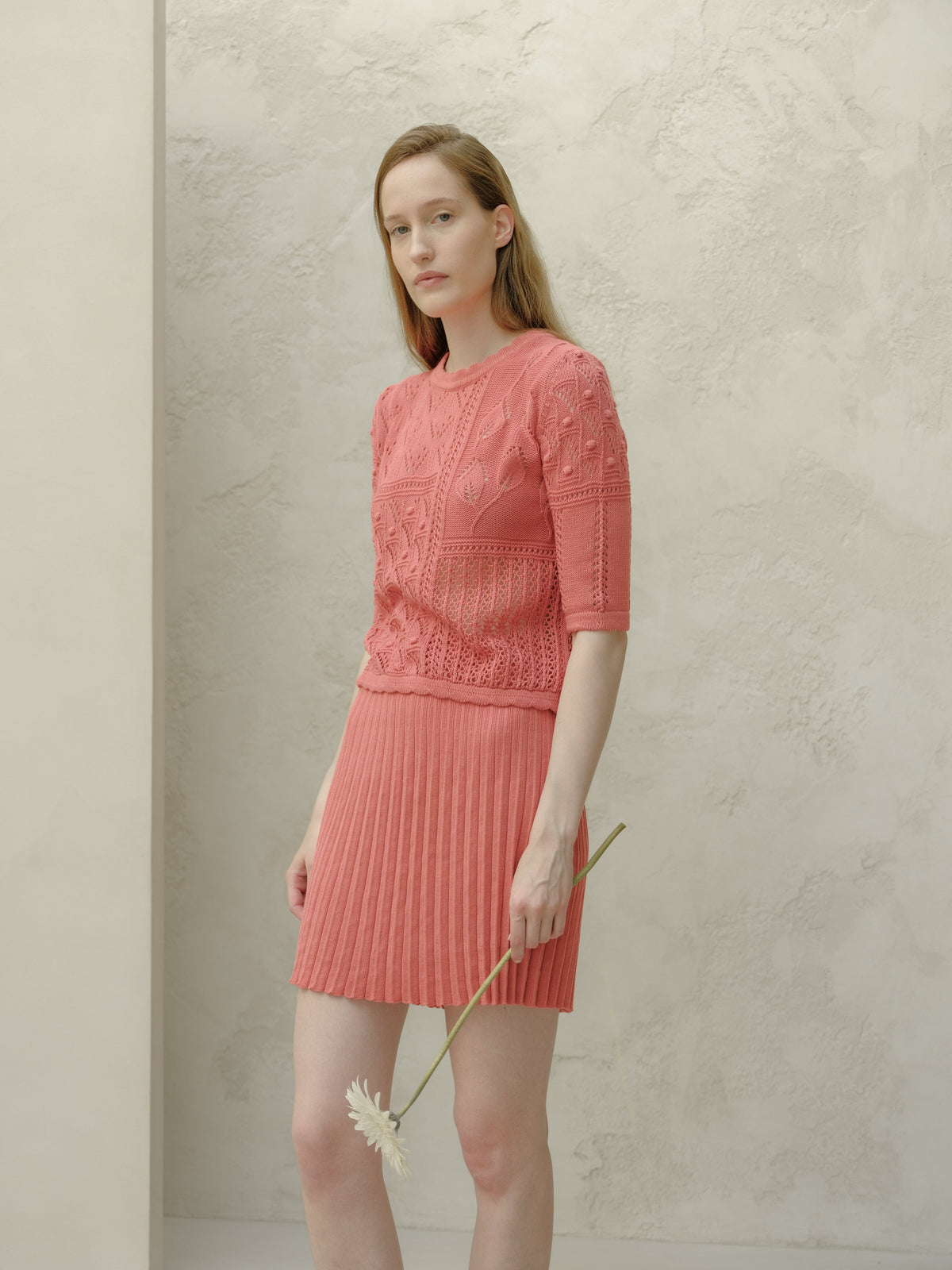 Model wearing pink Fine Cotton Plissé Mini Skirt with a matching knit top stands against a pale textured wall, holding a flower.