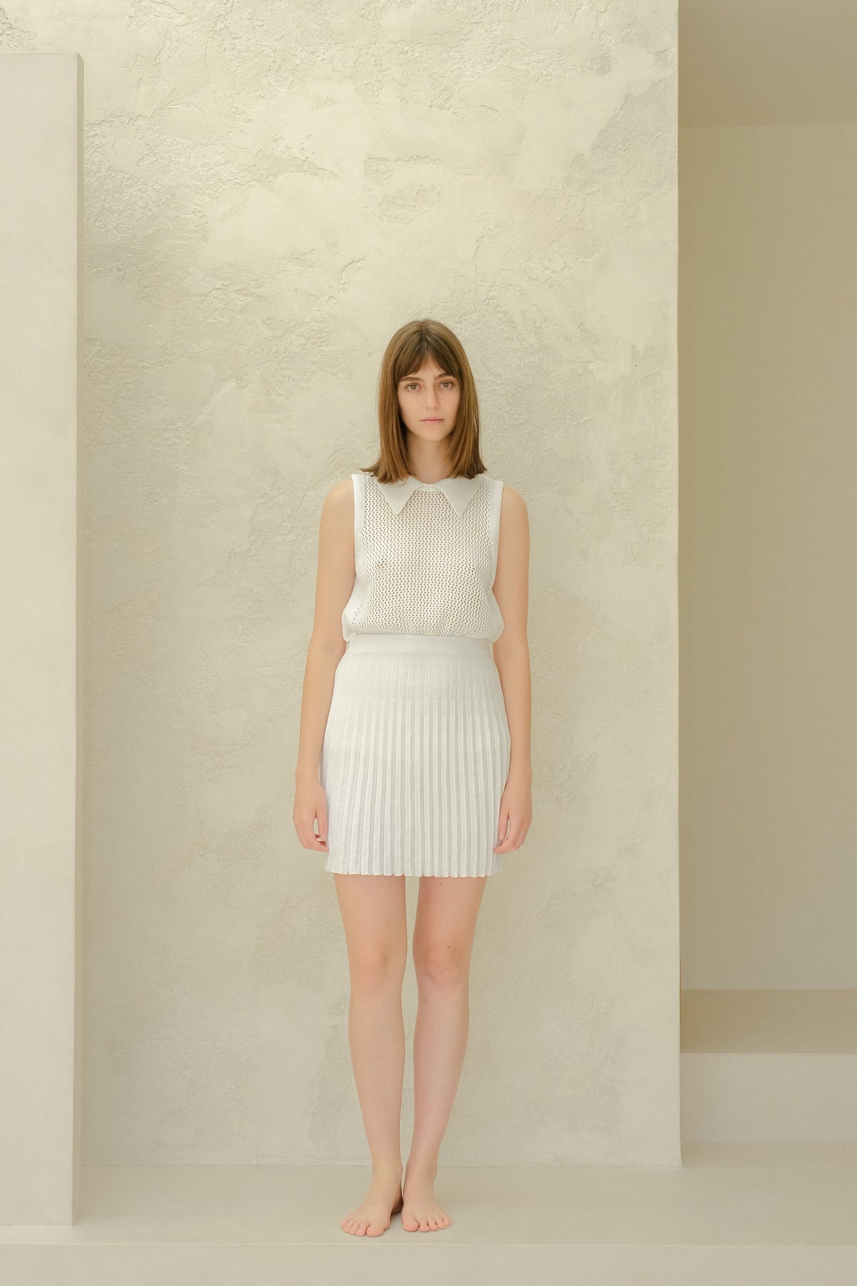 Full body of a model wearing a white mesh sleeveless top with a collared neckline and a white plissé mini skirt, barefoot.