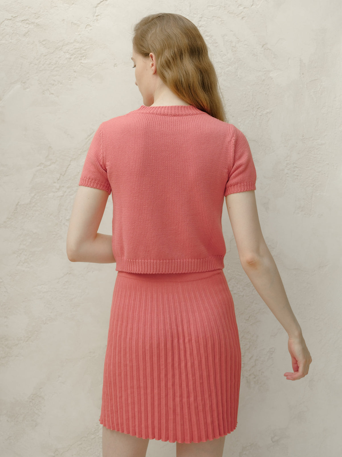 Back view of a model wearing a coral pink knitted top and matching pleated mini skirt