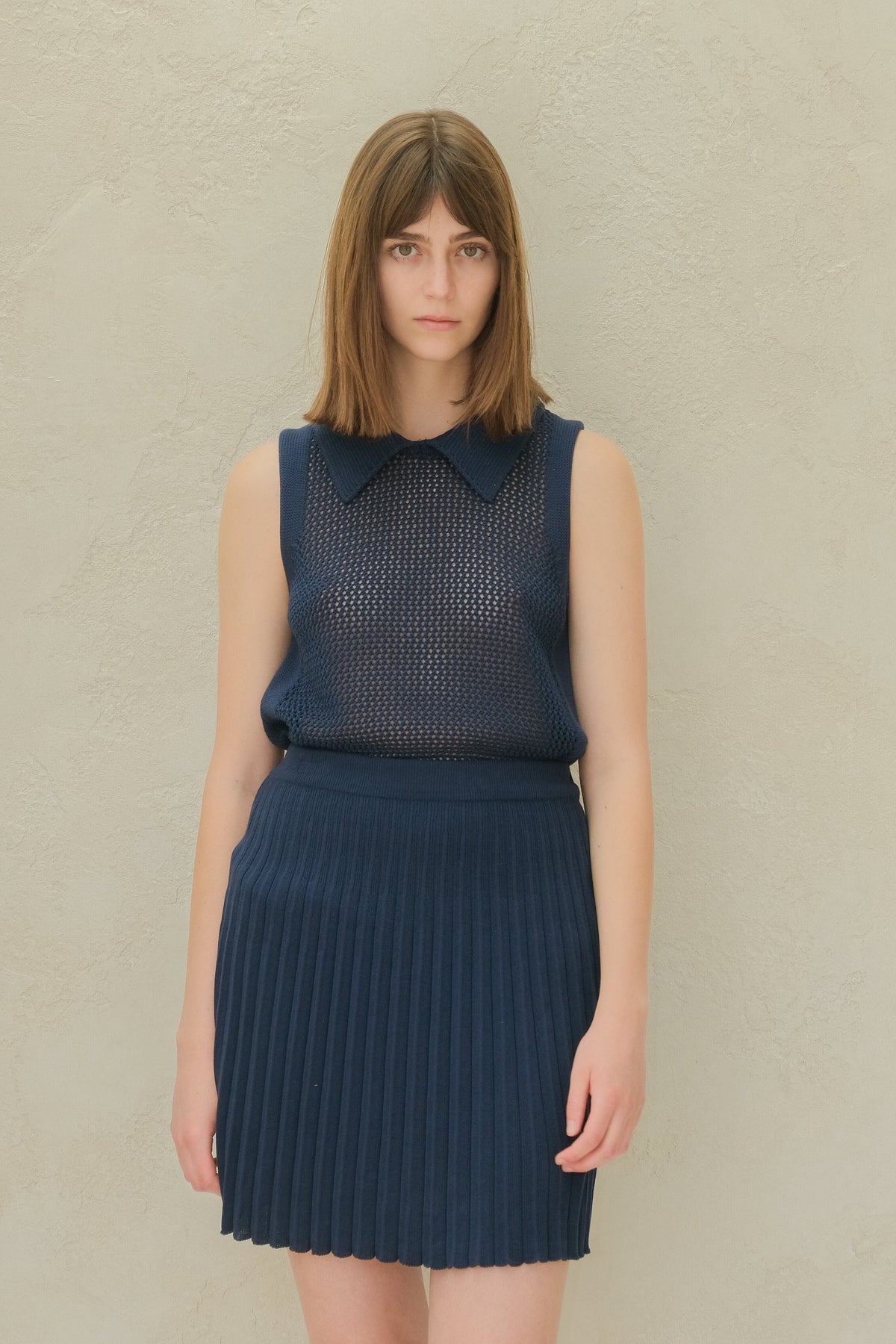 Navy sleeveless knit top with a mesh pattern and a pointed collar, paired with a navy pleated mini skirt.