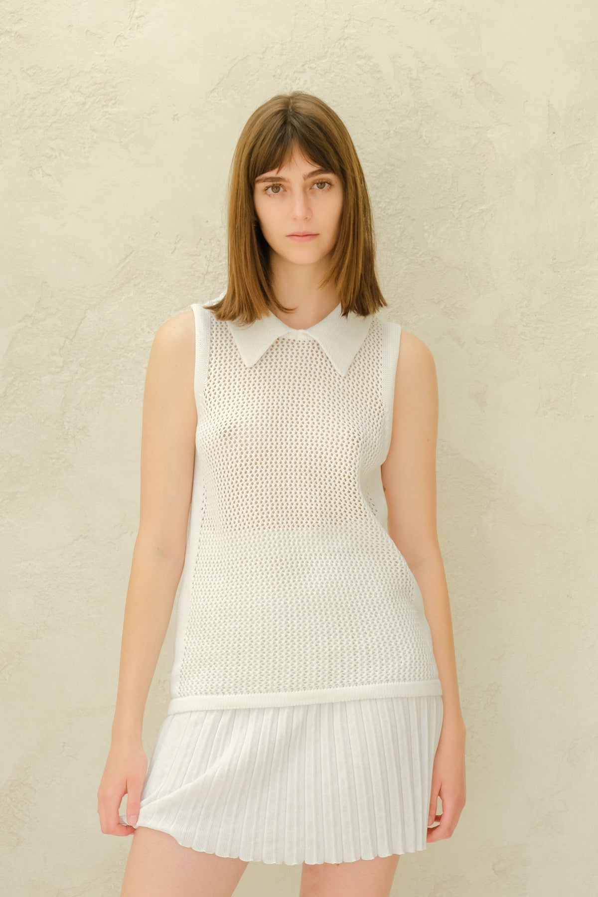 Model wearing a white mesh sleeveless top with a collared neckline and a white plissé mini skirt.