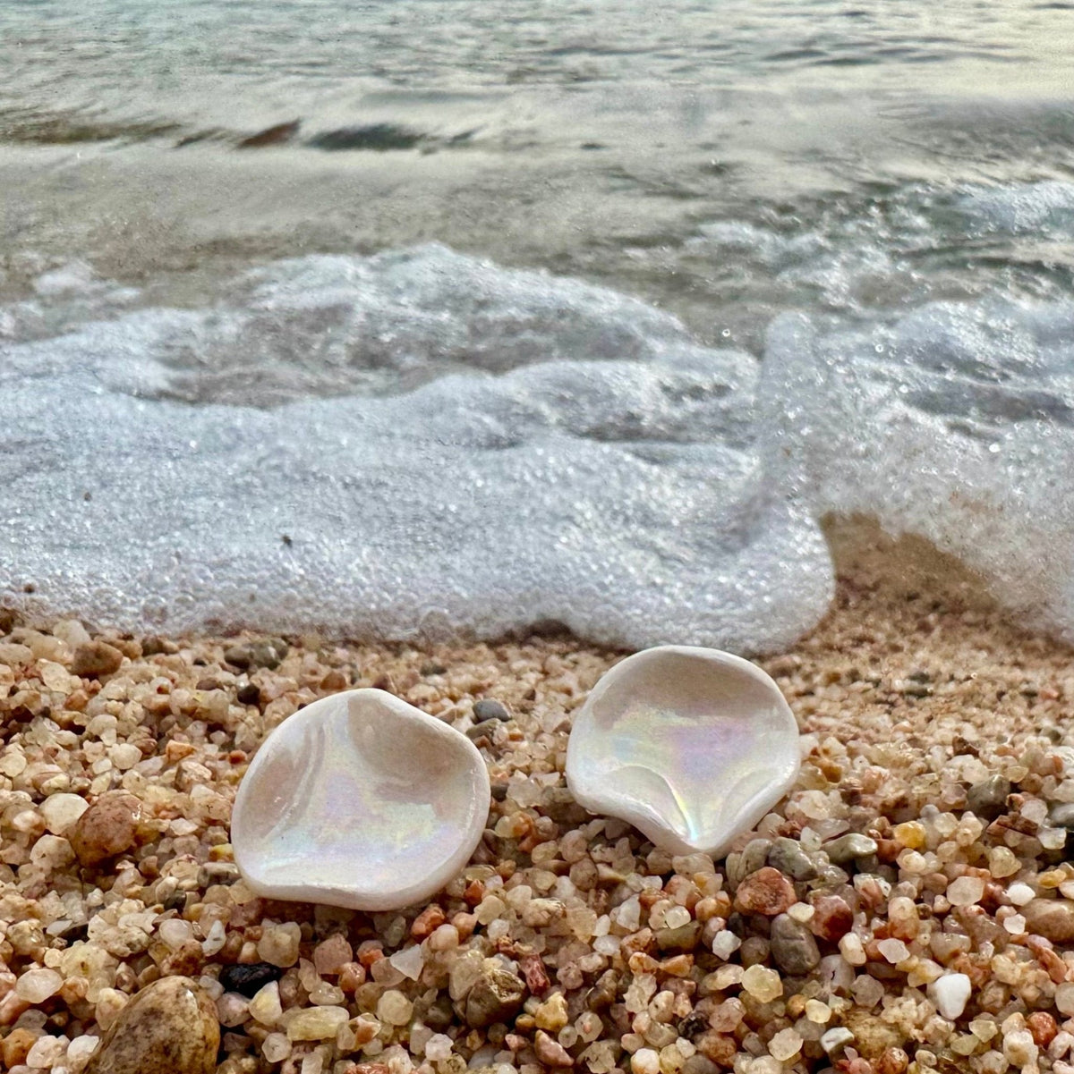 Two iridescent shell shaped earrings on a pebbled beach with waves in the background