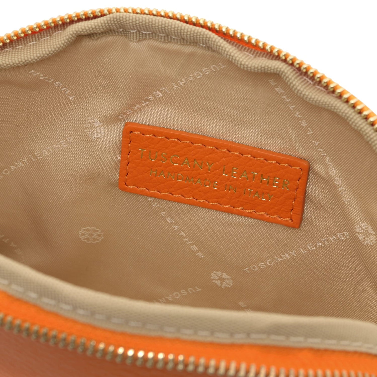 Gros plan of beige fabric doublure intérieure with embossed Tuscany Leather branding and stitched orange leather label inside toiletry case.