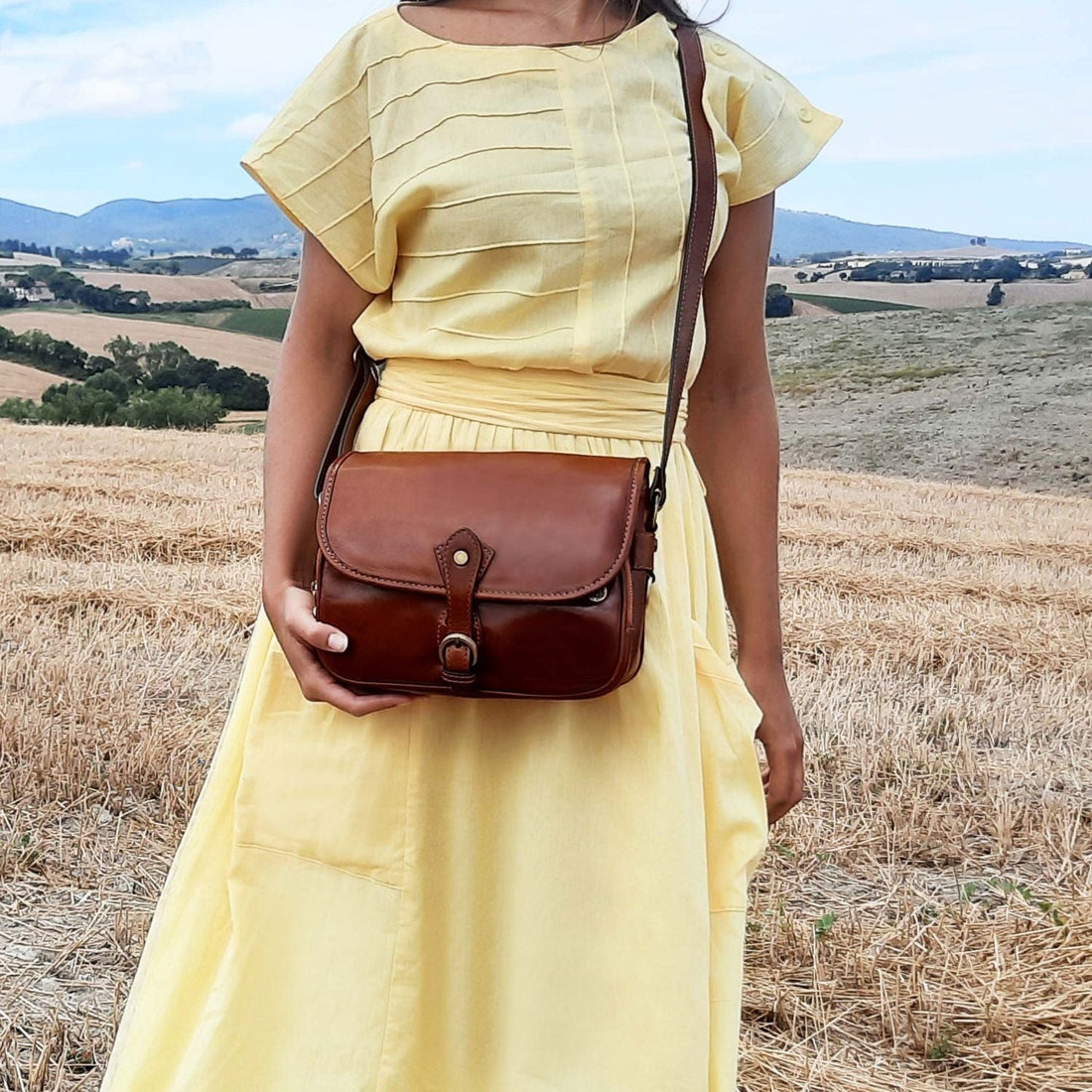 Woman in a pale yellow dress wearing a brown leather crossbody bag in a sunny countryside field.