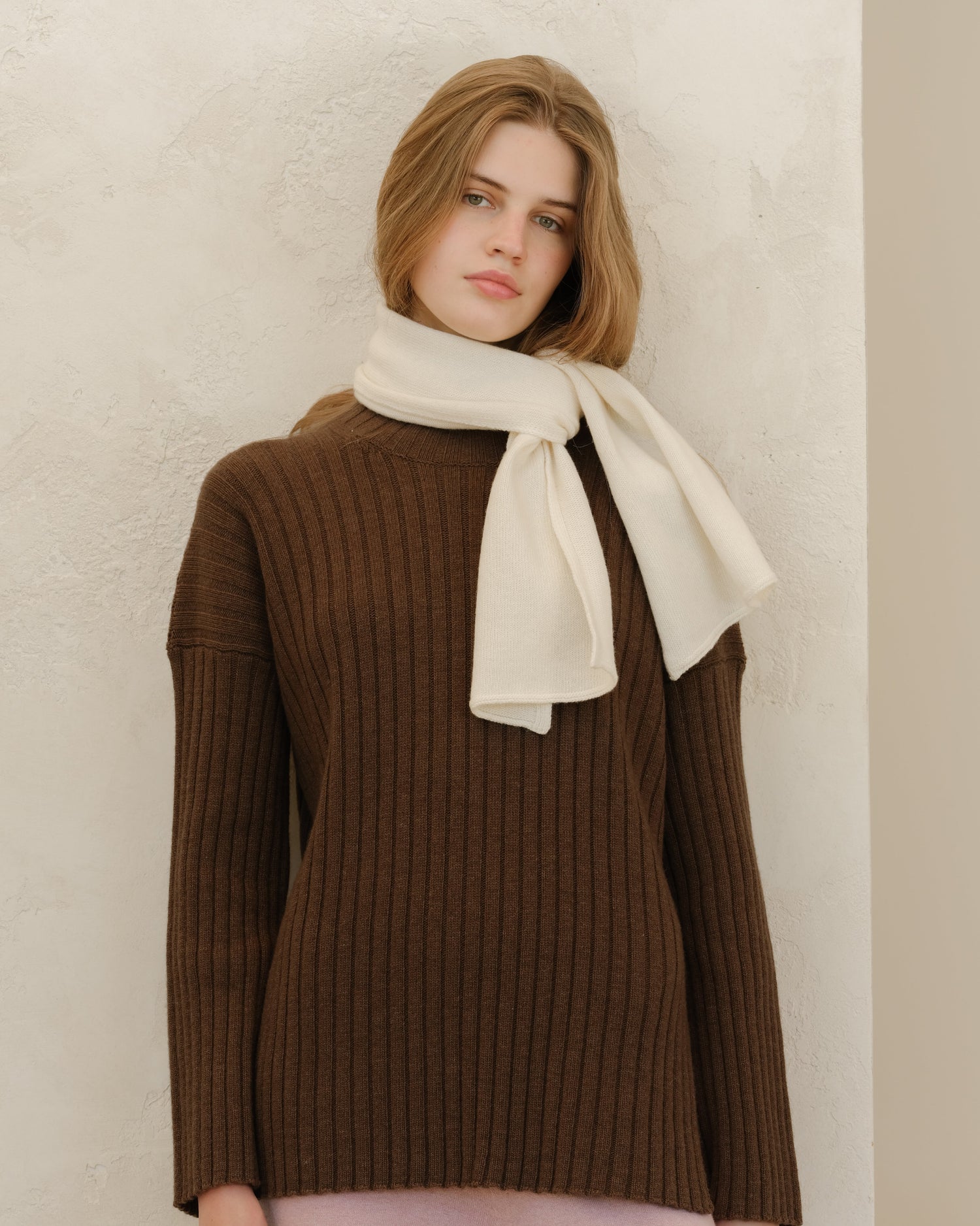Model in a cream blouse and beige tailored pants with a cream scarf beside an olive green handbag in a neutral setting from Autumn Winter 2025/26