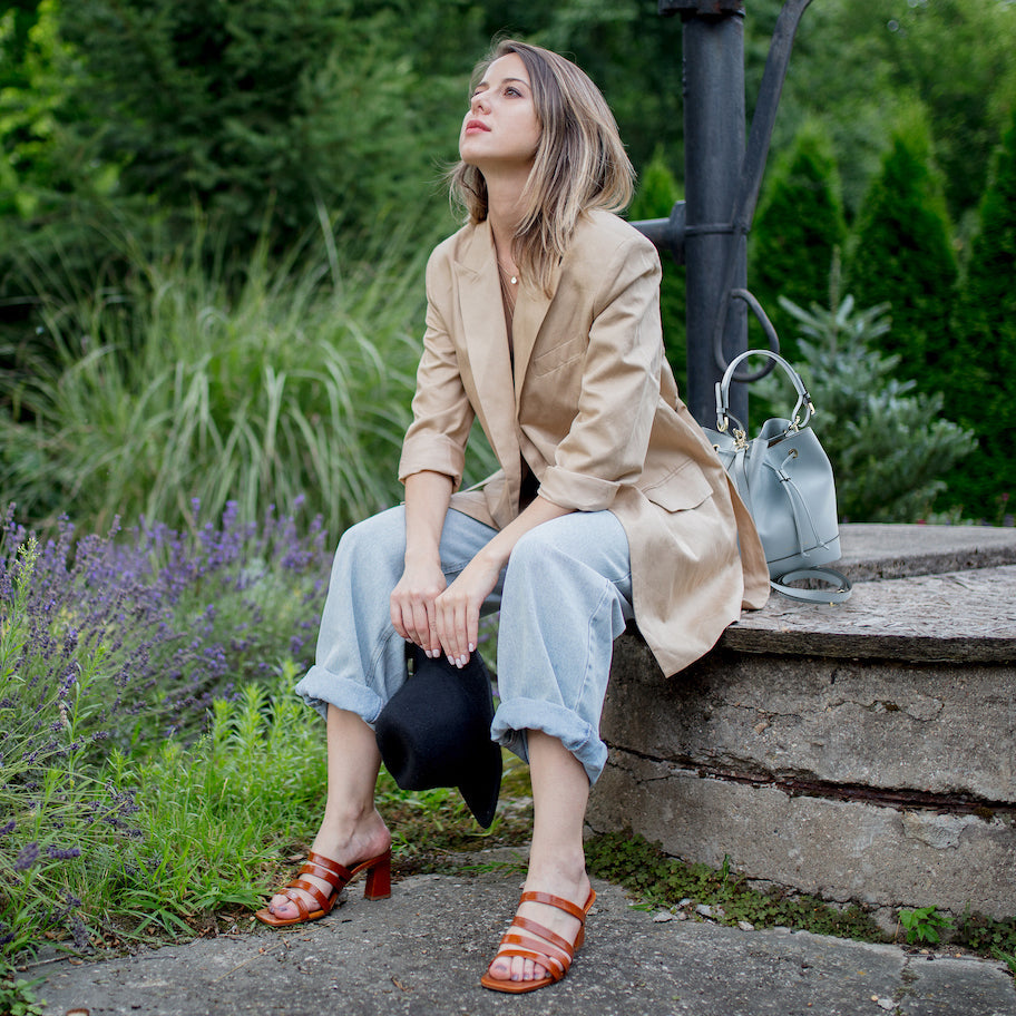 Woman in a beige blazer and light blue rolled jeans sits outdoors with a gray handbag beside her, wearing tan sandals.