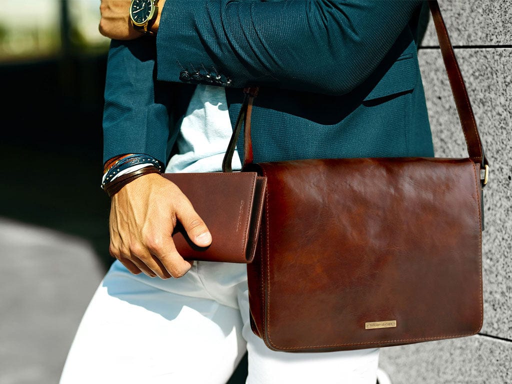 Man in a teal blazer holds a brown leather messenger bag and a matching wallet