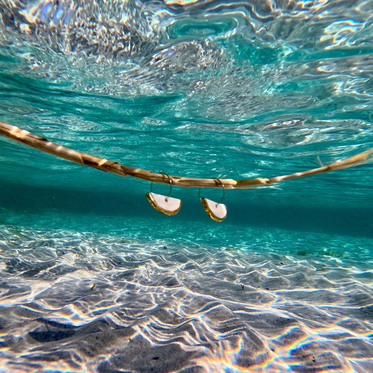 Gold hoop earrings with white crescent pendants hanging from a branch over turquoise water