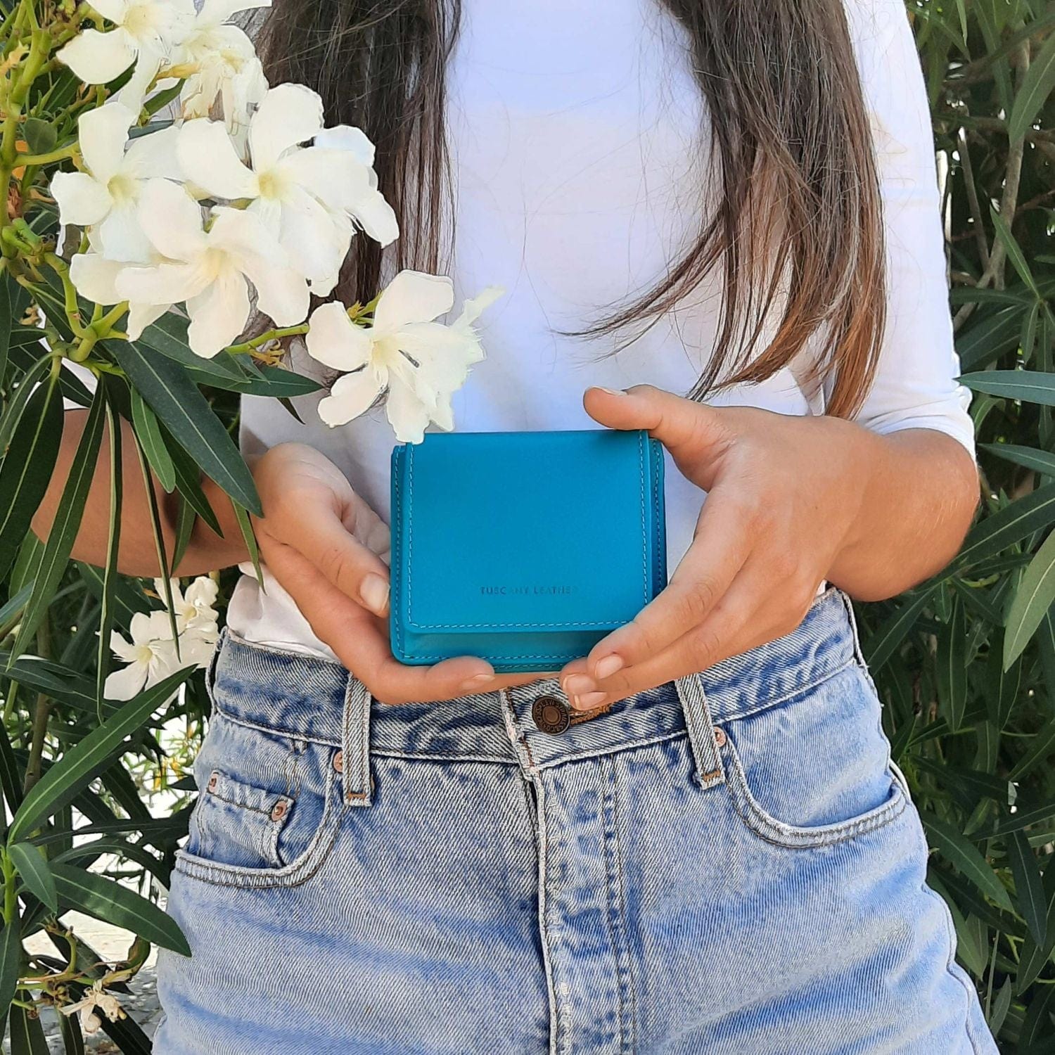 Woman in a white top and jeans holding a turquoise wallet outdoors with white flowers in the scene