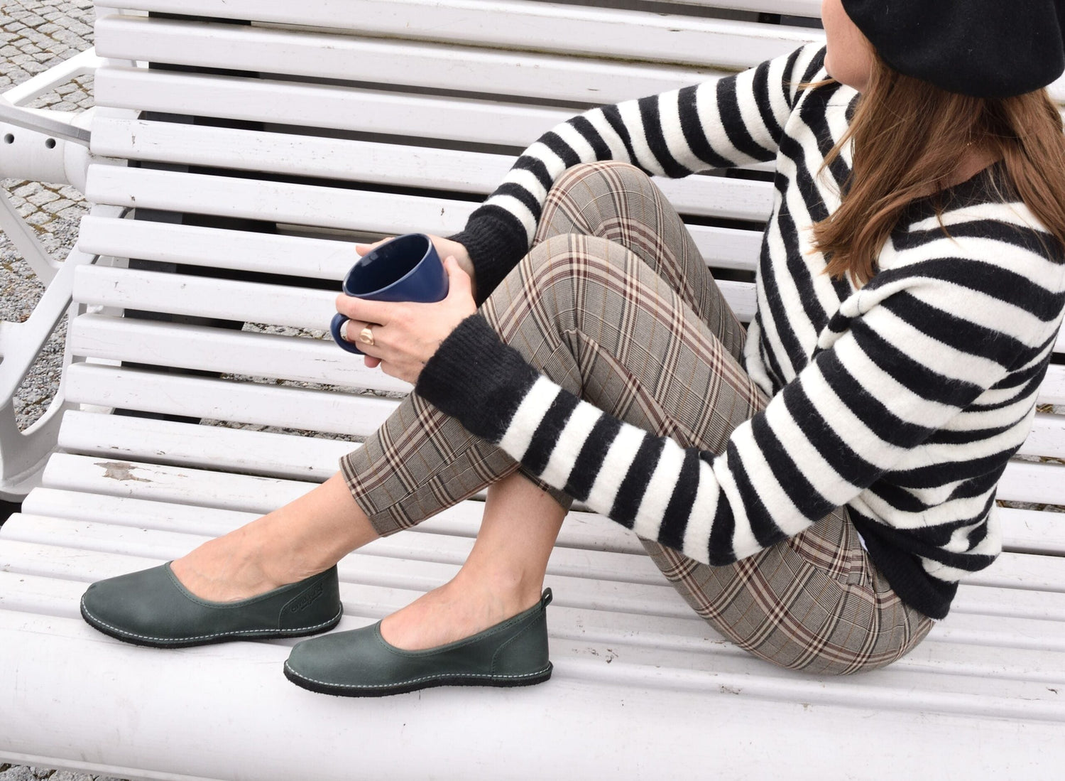 Woman sitting on a white bench wearing a black and white striped sweater and plaid pants with green leather ballet flats.
