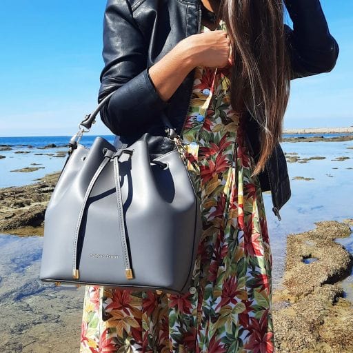 Woman wearing a floral dress and black jacket carries a gray Italian leather bucket bag by the sea