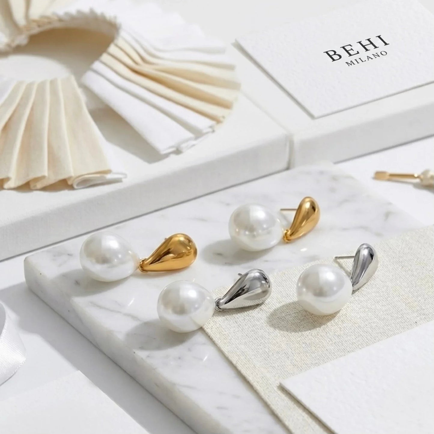Pearl and gold/silver earrings on a marble surface with 'BEHI MILANO' branding.