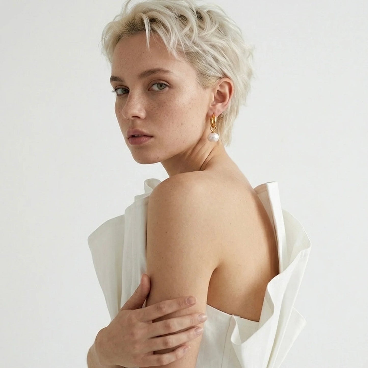 Woman with short blonde hair wearing gold and pearl earring in a white outfit against a plain background