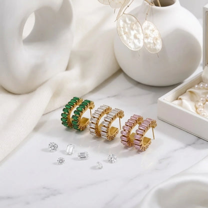 Three pairs of hoop earrings in green, gold, and pink on a marble surface with small gemstones.