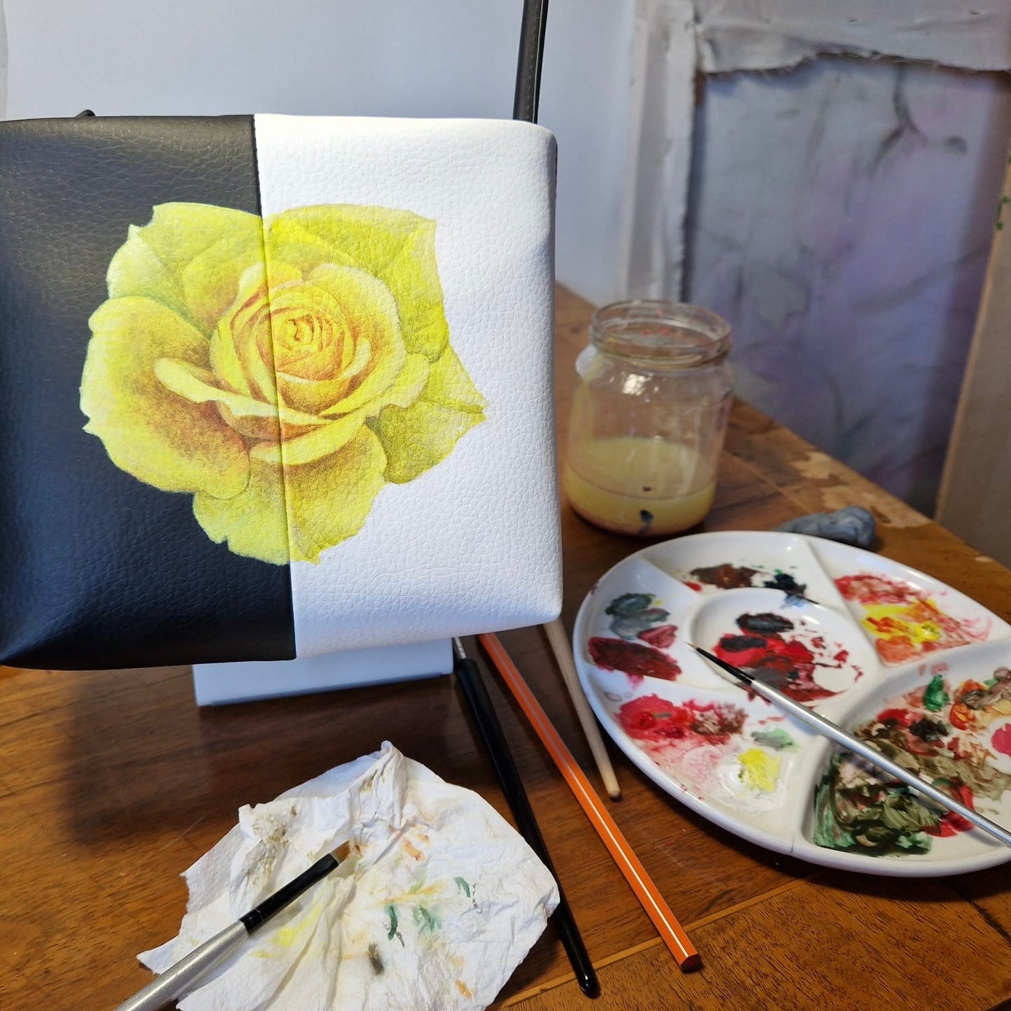 Leather handbag with a yellow rose painted on it, paint palette, and art supplies on a table.