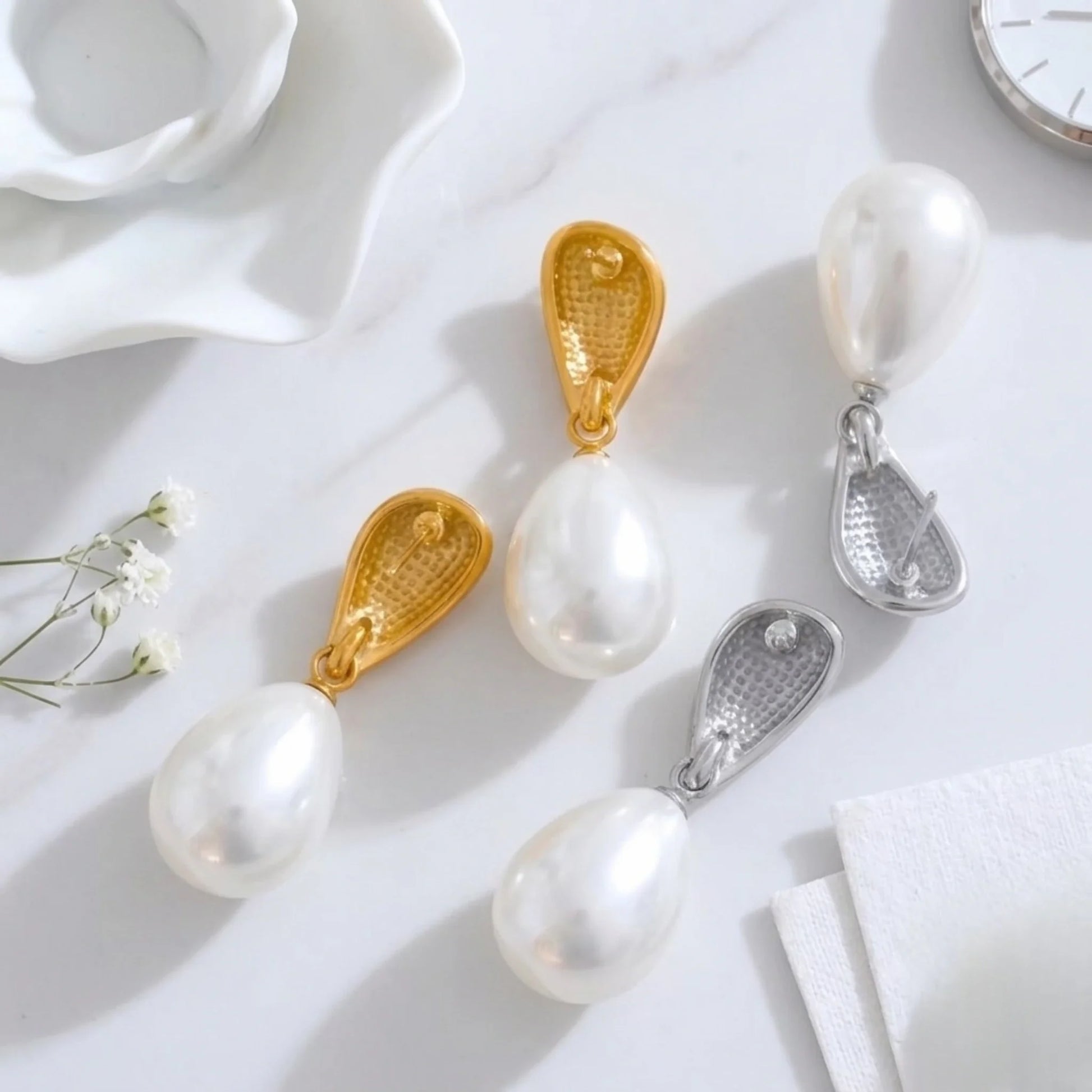 Back side of pearl earrings with gold and silver clasps on a white surface with flowers and a clock.
