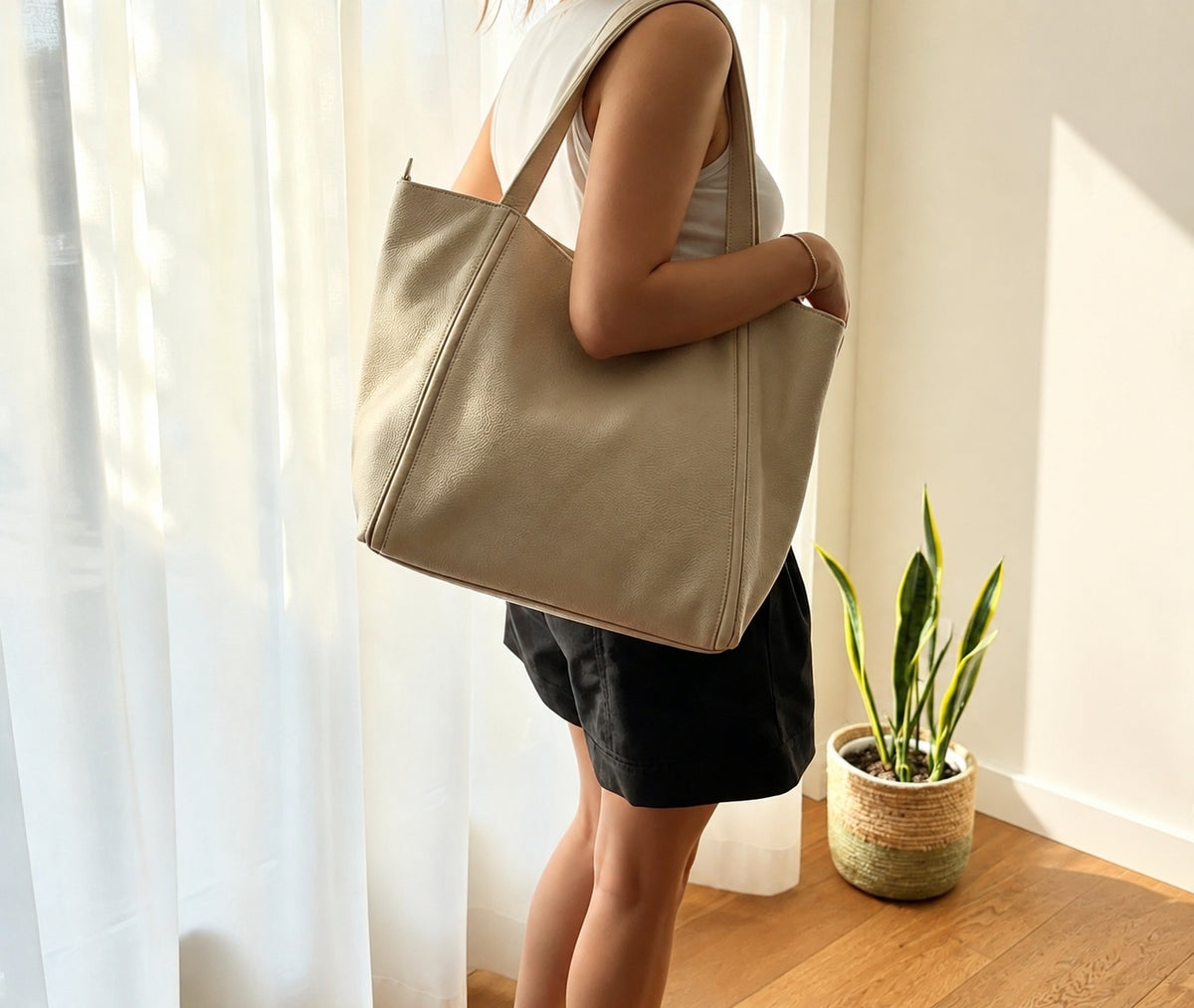 A woman wearing a white tank top and black shorts carries a large beige canvas tote bag over her shoulder in a brightly lit room with sheer curtains, a wooden floor, and a potted snake plant.