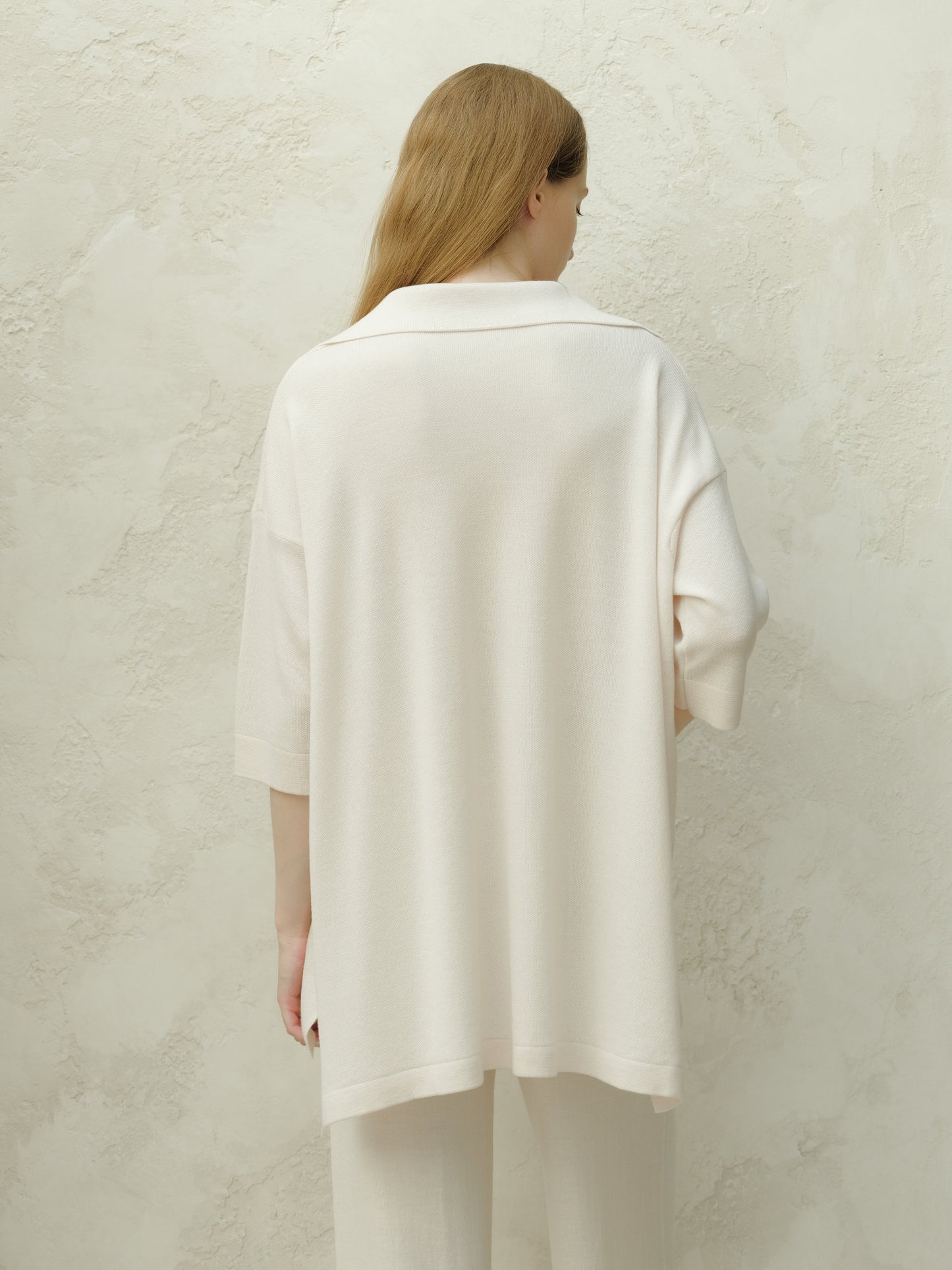 Back view of a model in a pale cream Merino Wool Maxi Polo with a loose drape against a textured wall