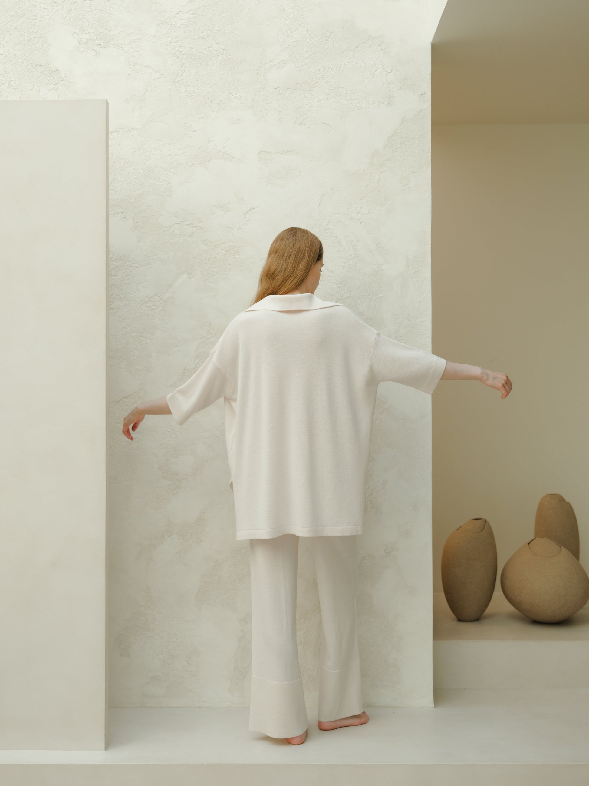 Back view of a model in an oversized white Merino wool maxi polo against a textured light wall