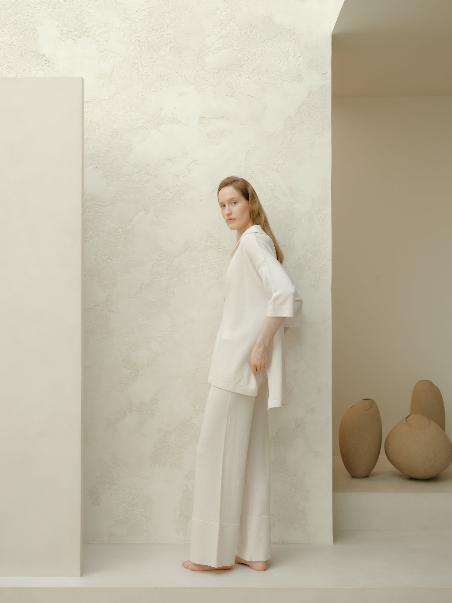 Model wearing a white Extra Fine Merino Wool Maxi Polo and matching wide-leg pants stands barefoot against a pale textured wall.