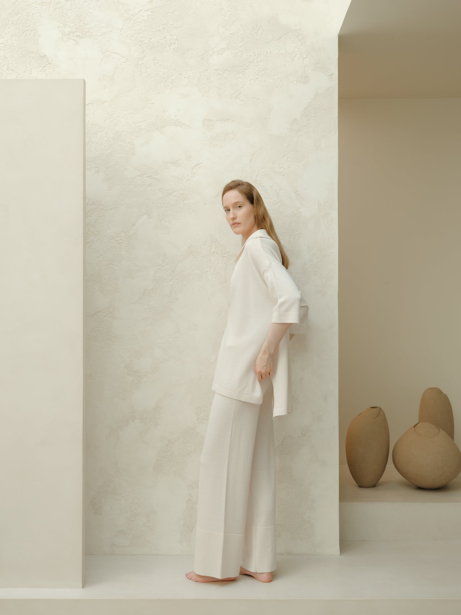 Model wearing a white Extra Fine Merino Wool Maxi Polo and matching wide-leg pants stands barefoot against a pale textured wall.