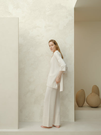 Model wearing a white Extra Fine Merino Wool Maxi Polo and matching wide-leg pants stands barefoot against a pale textured wall.