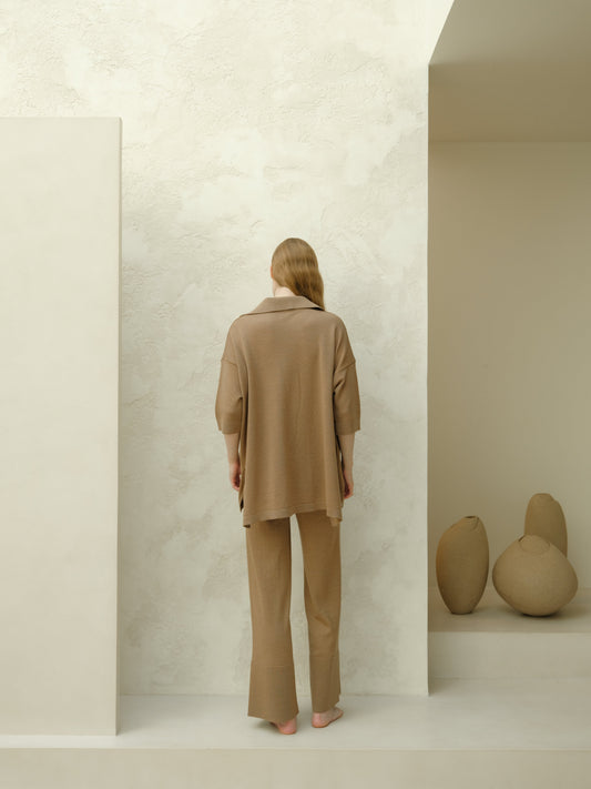 Back view of a woman in camel brown merino wool maxi polo set standing barefoot in a minimalist studio.