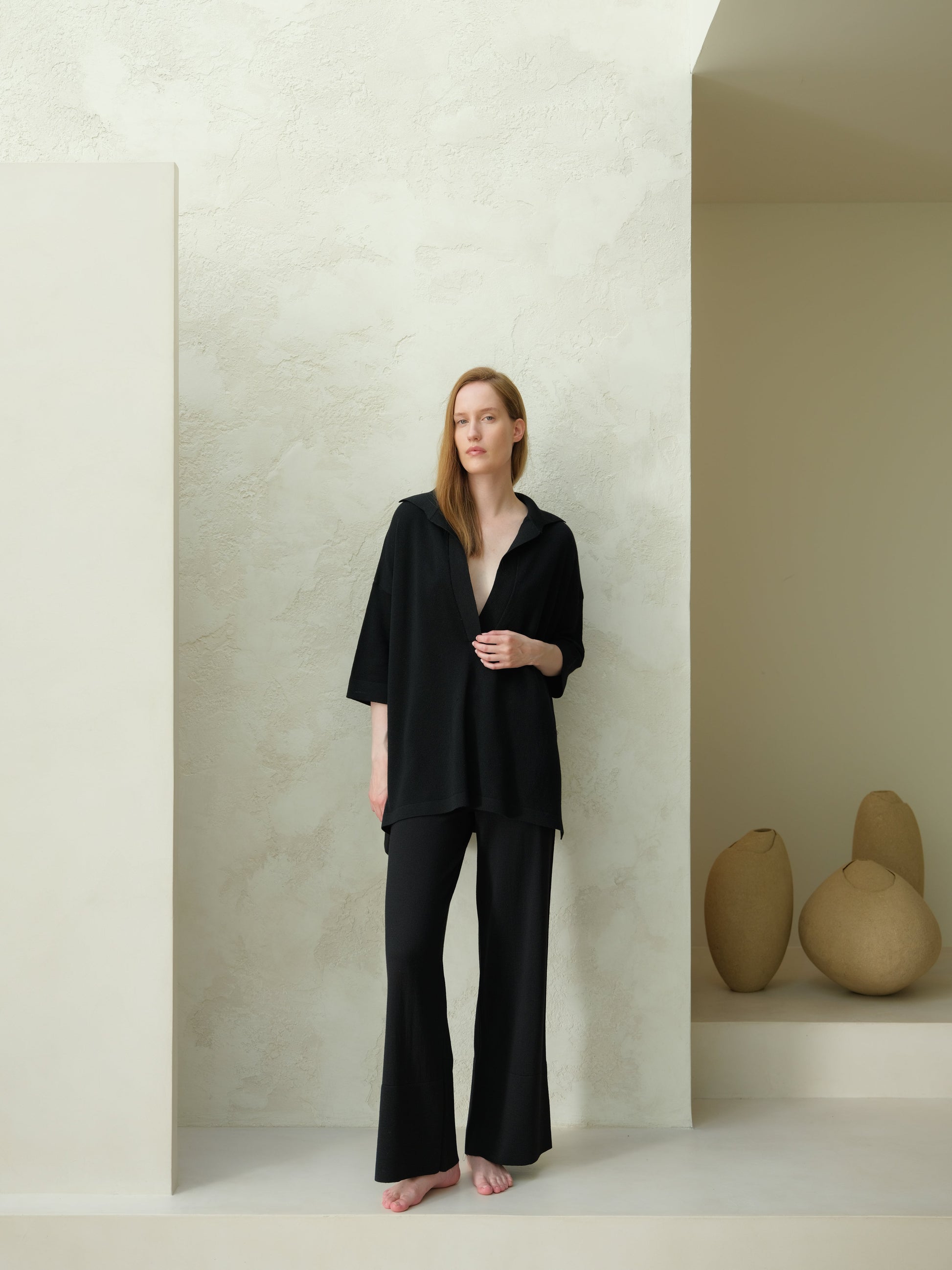 Full-body view of a model in a black Extra Fine Merino Wool Maxi Polo paired with wide-leg pants in a minimalist studio.