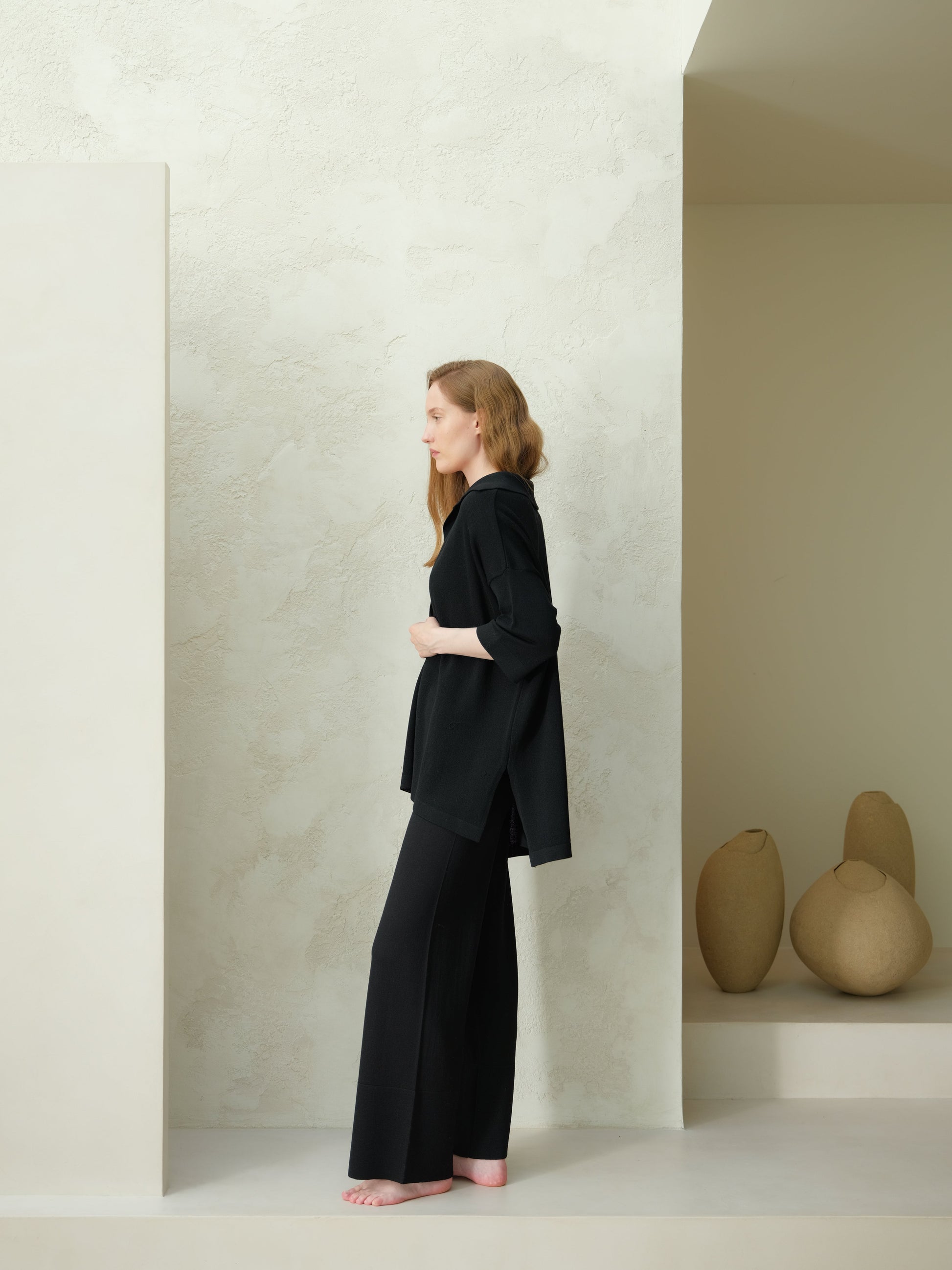 Profile view of a model wearing a black Extra Fine Merino Wool Maxi Polo with wide-leg trousers against a pale textured backdrop.